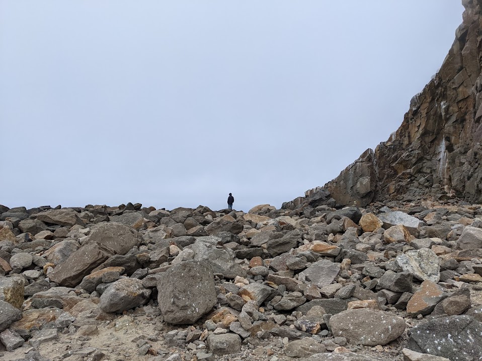 Another side of the hill. The rock of the hill blends into the ground, made of large boulders with a small person in the distance.