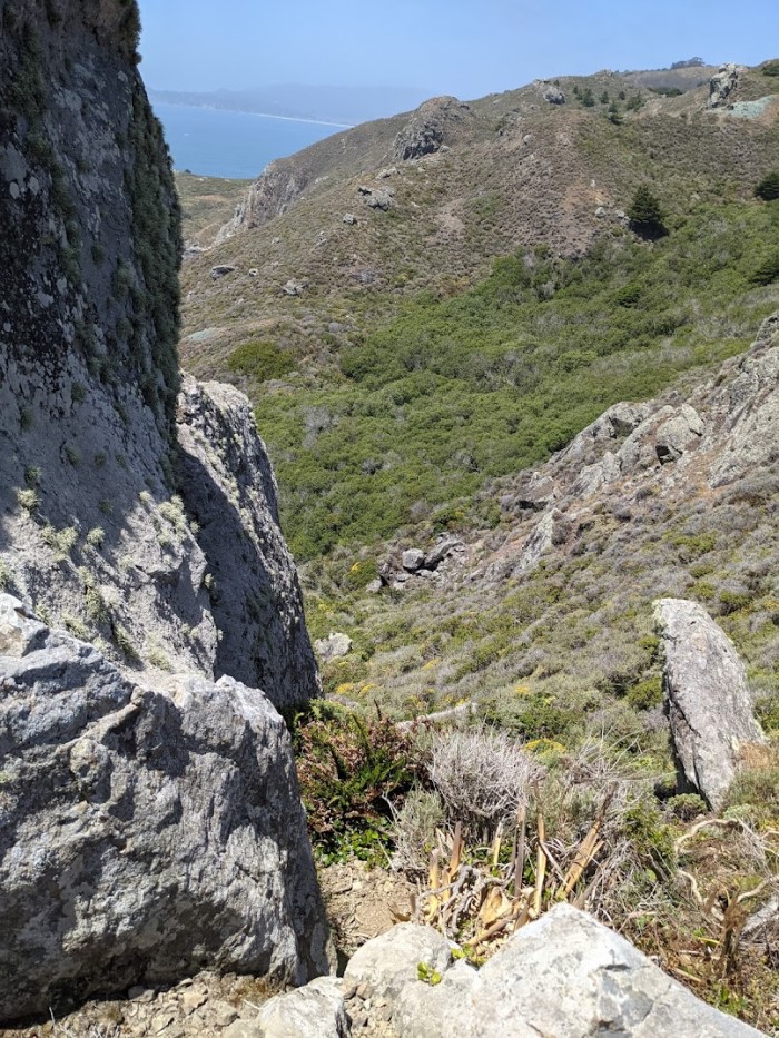 The side of a rocky cliff in the foreground, rocky hills with light vegetation behind. In the distance, the sea.