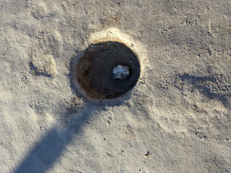 Close up of the ground on the path. Looks like packed dirt, a small hole a few inches across and deep shows water at the bottom, below the crust of salt, and a snowball-like clump in the middle.