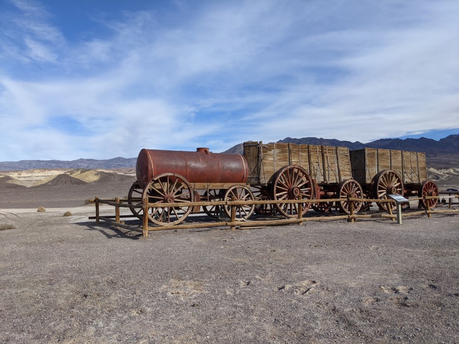 Picture of the cart from the other side. A giant rusted metal cylinder sits on a wooden cart with wooden wheels. Two wooden carts behind it with high wooden walls. Each bigger than a car. The front wheels are a lot bigger than the back for each.