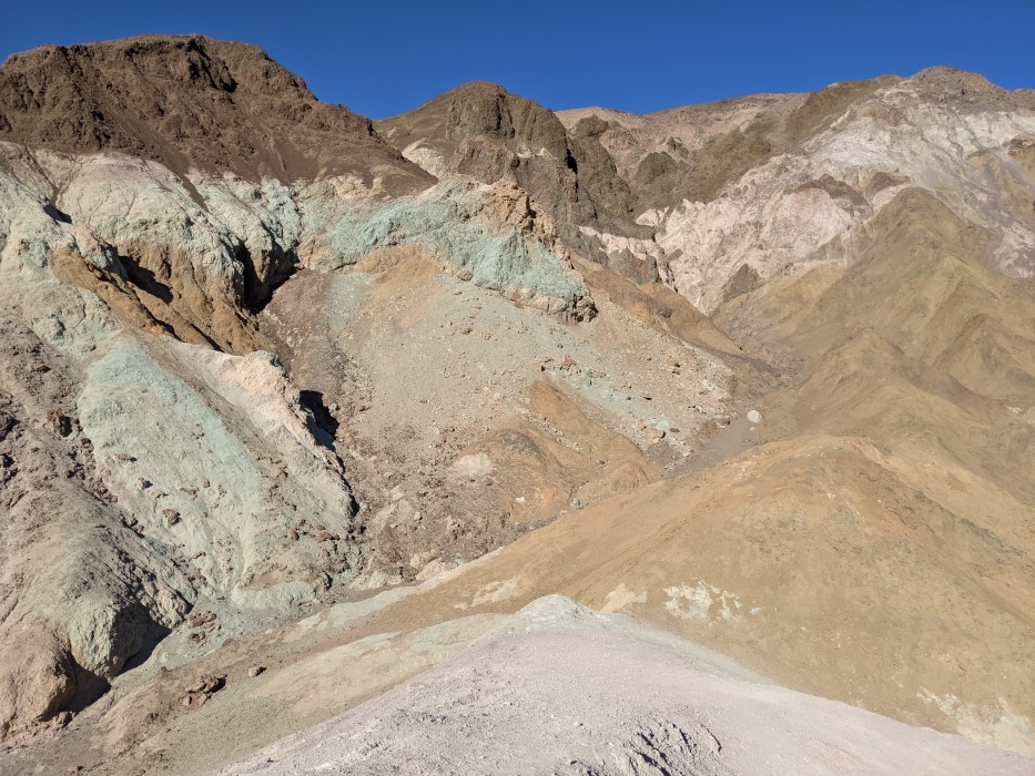 In the foreground, the purple hill the photographer is standing on. In the background, more hills, with subtle shades of green and pink on some of the rock faces. Everything is dry and without vegetation.