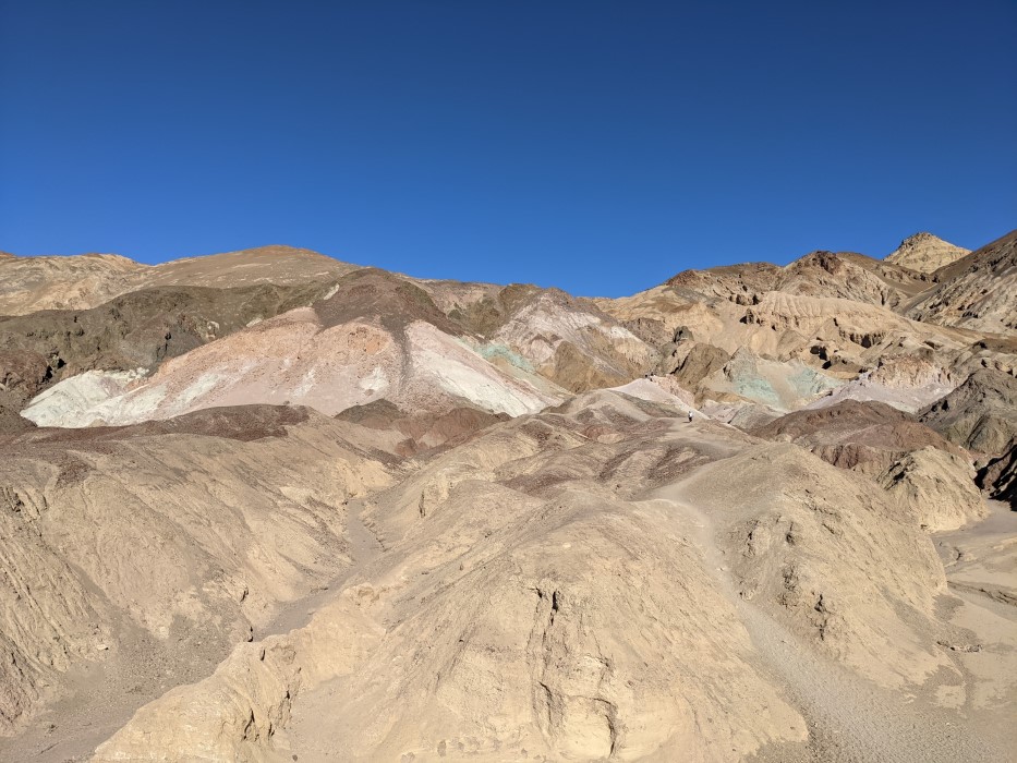 In the distance, the painted hills shown earlier, but with many small hills in between. A hiker closer to the hills is tiny.